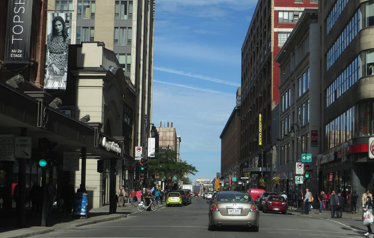 Blick auf die Rue Sainte-Catherine in Montréal mit Schaufenstern, Einkaufenden und markanten Leuchtreklamen auf einer belebten Einkaufsstraße unter klarem Himmel.