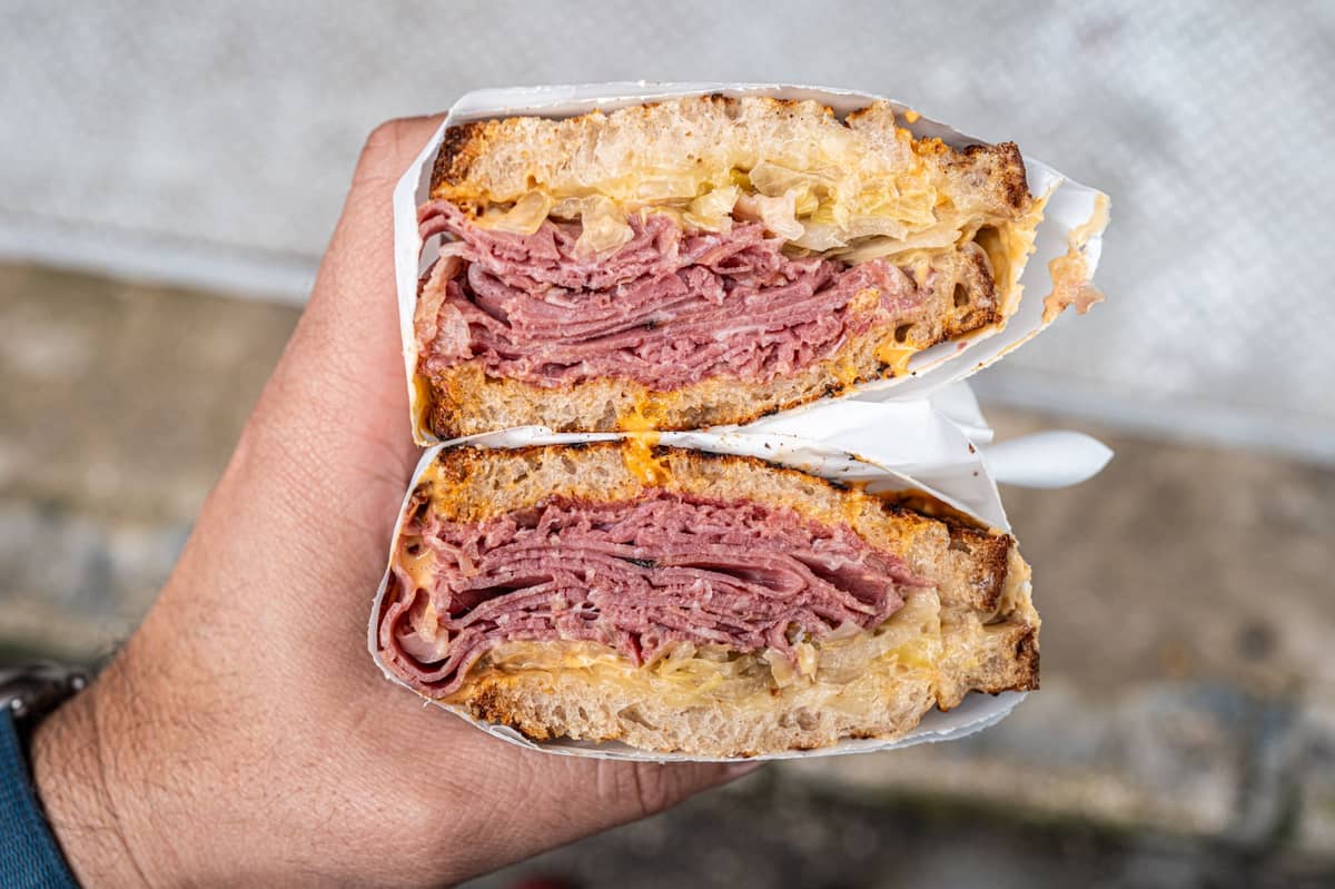 Nahaufnahme einer Hand, die ein Montrealer Smoked-Meat-Sandwich hält – dicht belegt mit gewürztem Rindfleisch auf Roggenbrot.