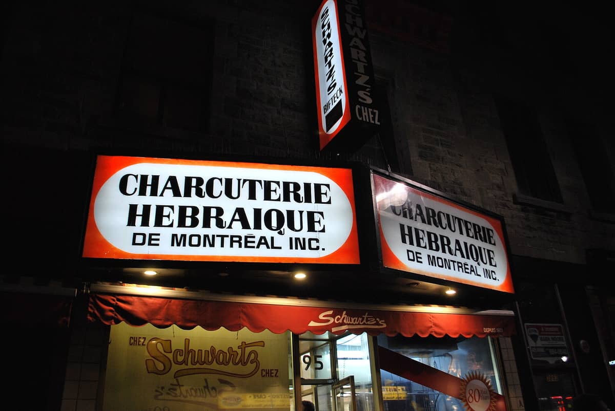 Foto notturna dell'iconica facciata della salumeria Schwartz's a Montreal con insegne luminose rosso e bianco e ingresso in vetro.