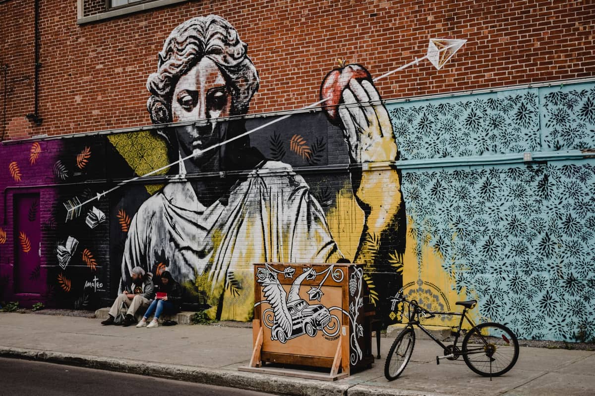 Duży mural na ceglanych budynku w Montrealu z siedzącymi w pobliżu ludźmi, ukazujący żywą sztukę uliczną i atmosferę festiwalu.