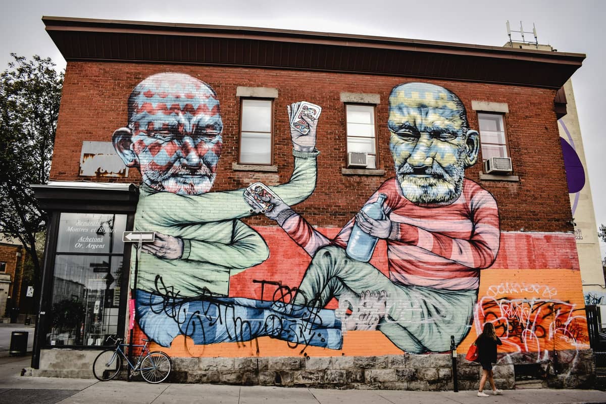 Farbenfrohes Wandgemälde in Montreal mit zwei großen sitzenden Figuren, die miteinander trinken – gemalt auf die Backsteinwand eines Gebäudes in einem städtischen Viertel.