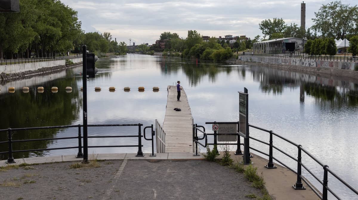 Widok na Lachine Canal w Montrealu z pływającym pomostem, balustradami i zielenią po obu stronach pod zachmurzonym niebem.