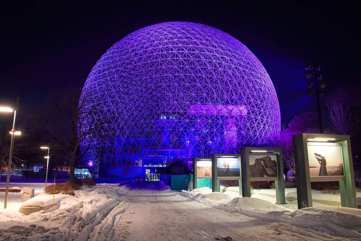 Nächtliche Winterszene an Montreals Biosphäre, in violettes Licht getaucht, mit Schnee auf dem Boden und Kunstinstallationen, die zur Kuppel führen.