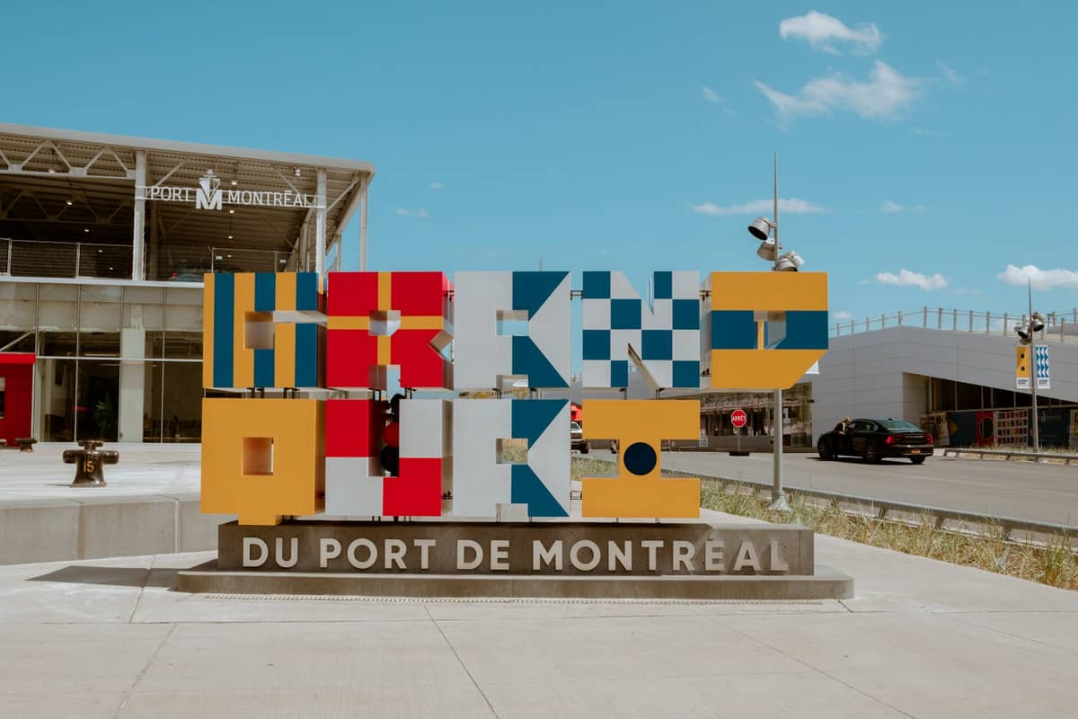 Kolorowy, nowoczesny znak z napisem 'Grand Quai du Port de Montréal' przed budynkami Starego Portu w słoneczny dzień.