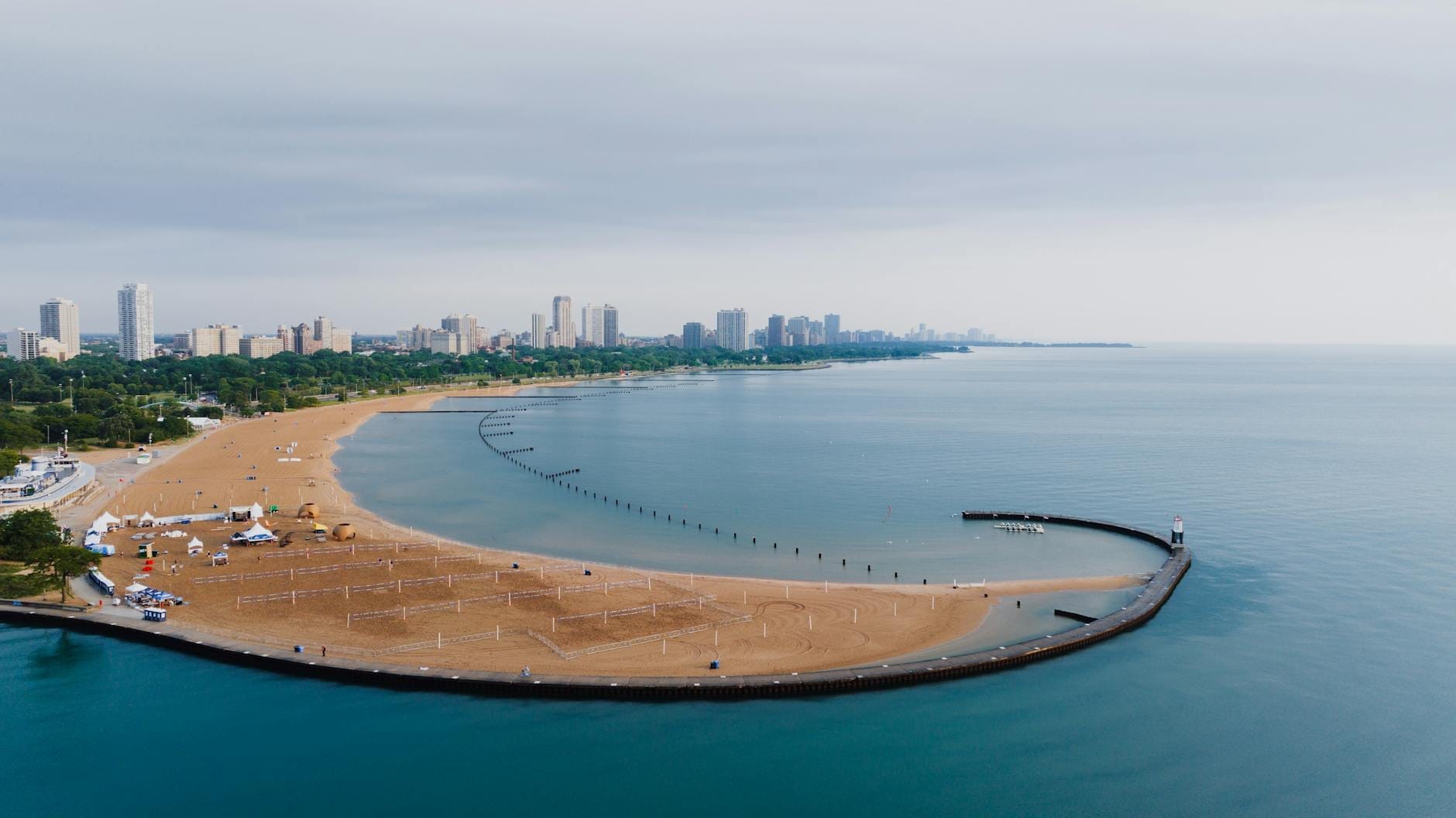 Un'ampia spiaggia sabbiosa con campi da beach volley, acqua calma e trasparente, e lo skyline di Chicago sullo sfondo sotto un cielo nuvoloso.
