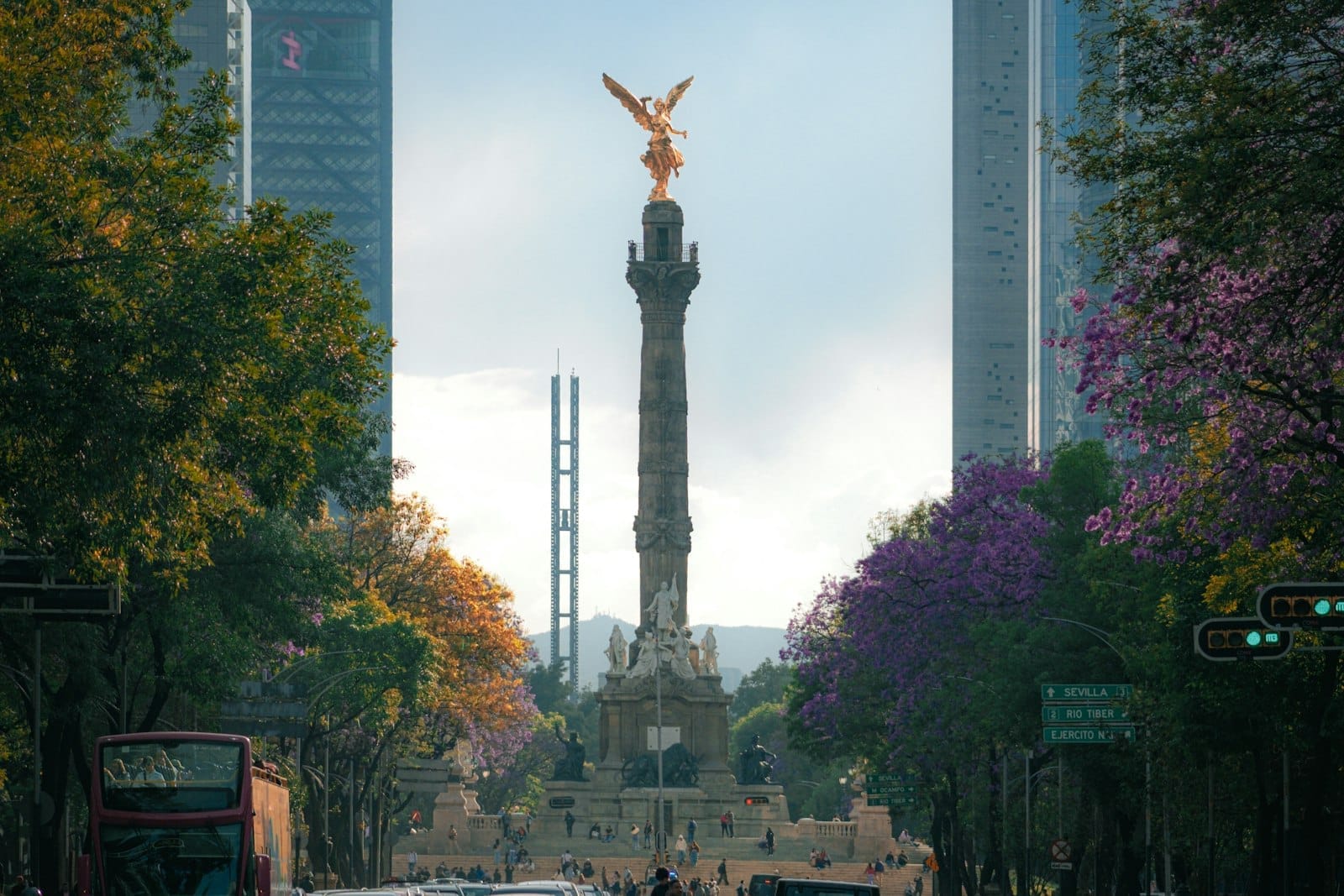 Il monumento dell'Angelo dell'Indipendenza si erge al centro del Paseo de la Reforma, incorniciato da edifici moderni e alberi con fiori viola sotto un cielo spettacolare.