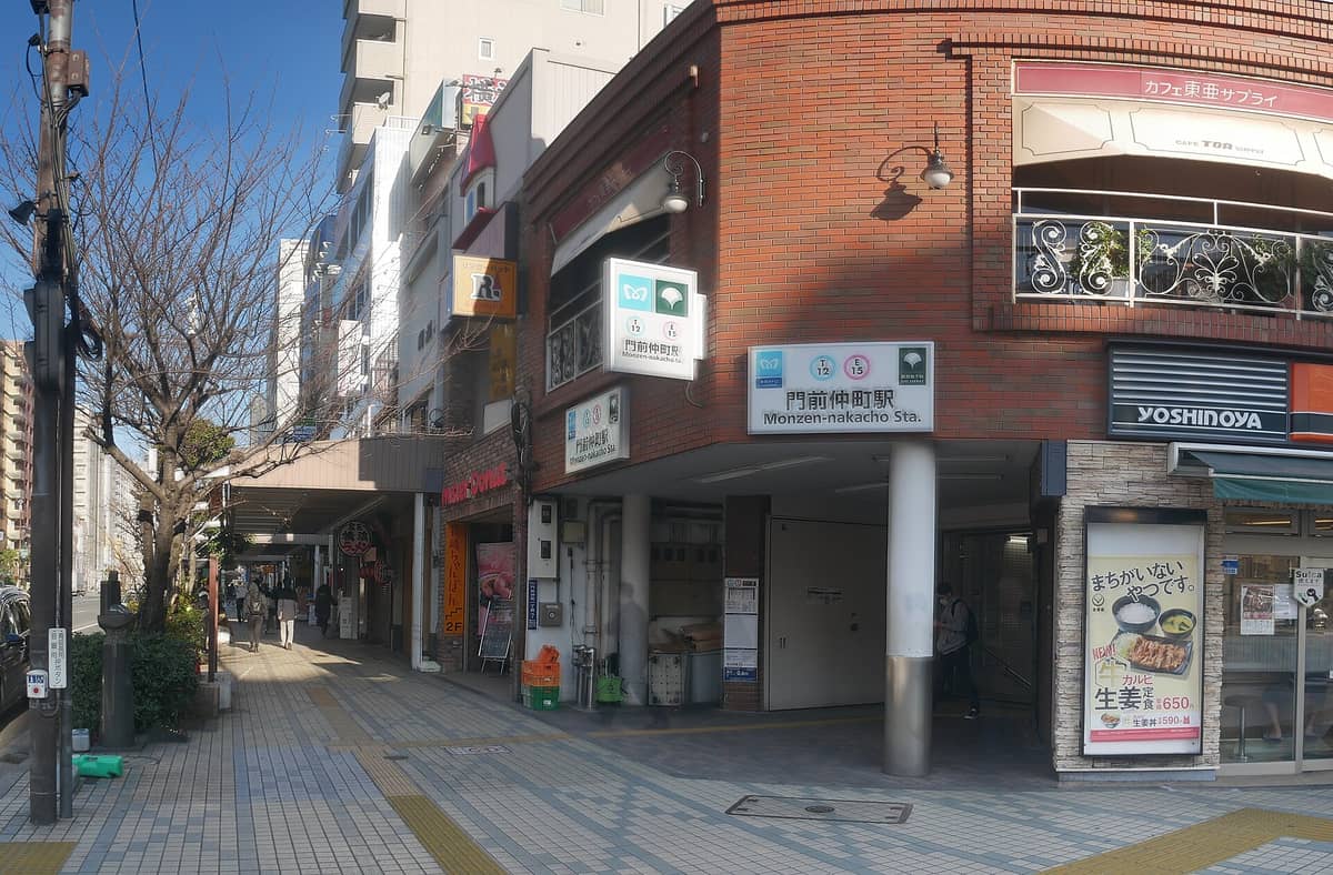 Street view of the entrance to Monzen-nakacho Station in Tokyo, with signage clearly showing the station name and a mix of local shops and city sidewalk.