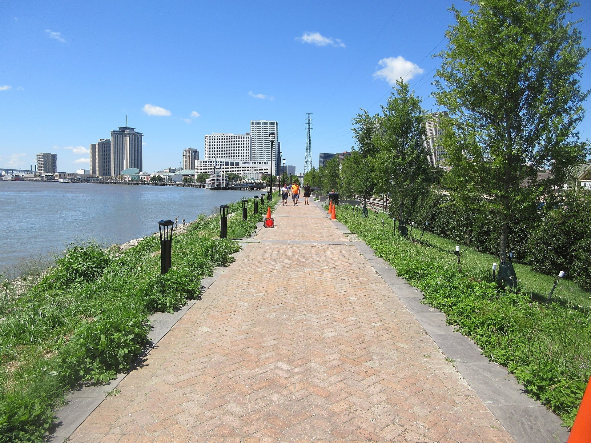 Passeggiata Moon Walk pavimentata in mattoni con persone che camminano, alberi verdi e vista sullo skyline del centro di New Orleans lungo il fiume Mississippi.