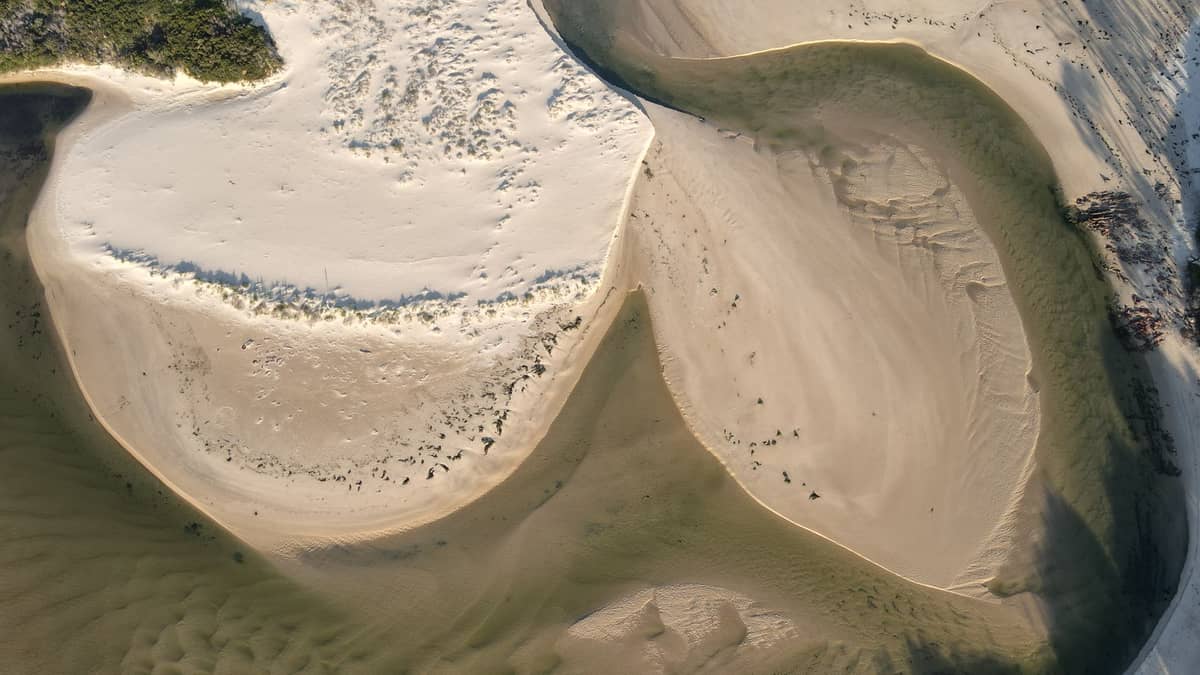 Vista aérea de la costa arenosa y las dunas que se encuentran con las aguas poco profundas de color azul verdoso, mostrando un paisaje natural virgen típico de Moreton Island.