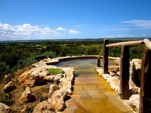 Geothermisches Hügelbecken der Peninsula Hot Springs auf der Mornington Peninsula, mit hellem Steinrand, Holzgeländer und Buschland bis zum Horizont unter blauem Himmel.