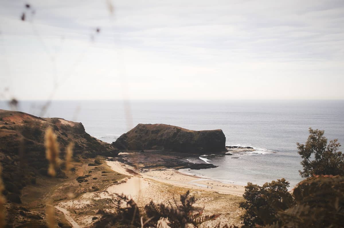 Afloramiento rocoso y playa de arena en la Península de Mornington bajo una luz diurna suave, con olas tranquilas del océano y vegetación nativa en primer plano.