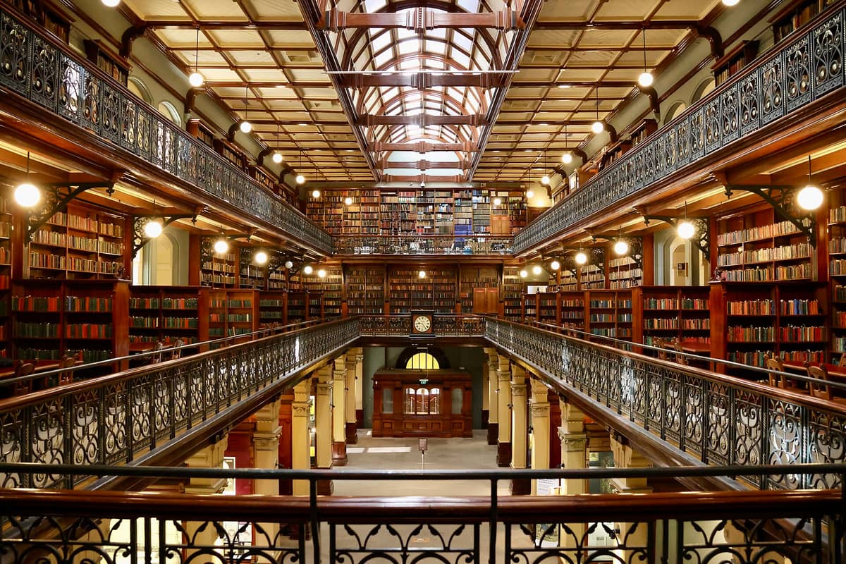 Interior of the Mortlock Wing, State Library of South Australia, featuring ornate cast-iron galleries, skylit ceiling, and shelves filled with books.