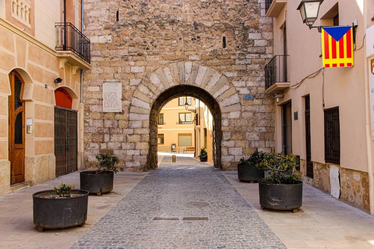 Steinbogeneingang zur Altstadt von Alcúdia mit historischen Gebäuden und einer mallorquinischen Flagge, der das Dorferbe Nordmallorquas widerspiegelt.