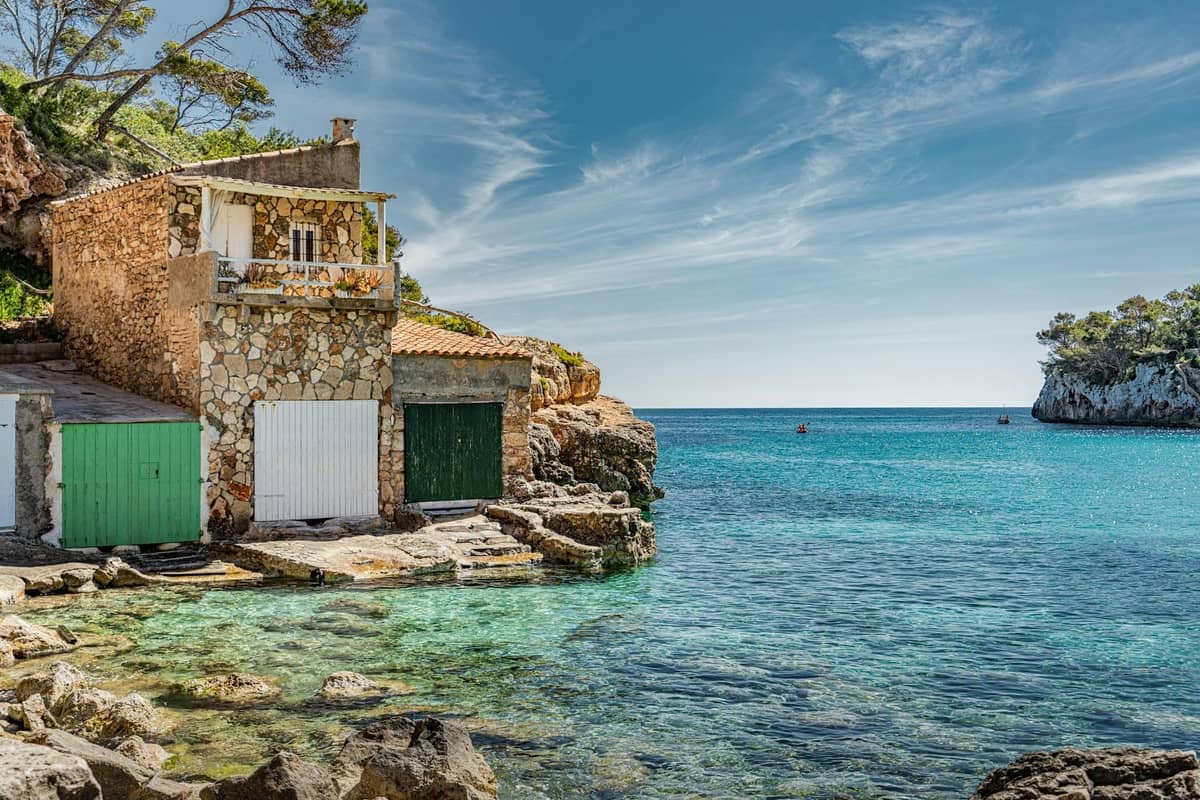 Traditionelle Steinbootshäuser an einer türkisfarbenen Bucht an der Südostküste von Mallorca, mit felsigem Ufer, klarem Wasser und malerischem Blick auf das Meer.