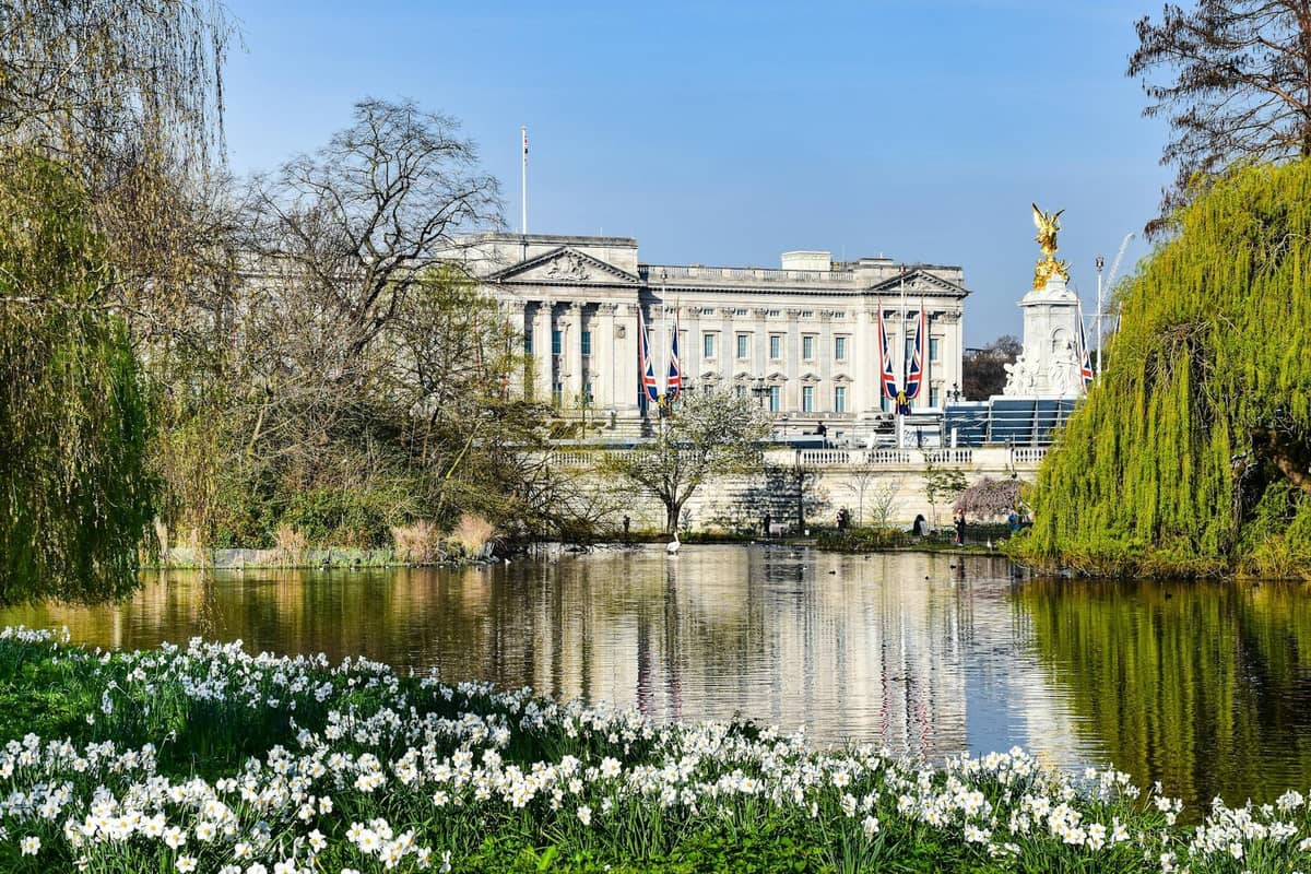 Buckingham Palace e o Victoria Memorial vistos de um lago cênico com flores primaverais e árvores frondosas em um parque no centro de Londres.