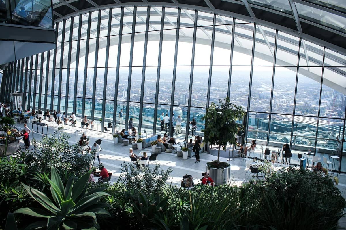 Interior do Sky Garden em Londres com grandes janelas de vidro, plantas exuberantes e visitantes aproveitando vistas panorâmicas do horizonte da cidade.