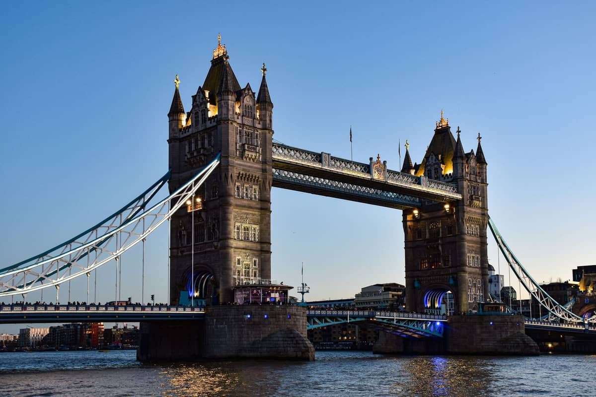 Tower Bridge em Londres ao entardecer, totalmente iluminada, com céu azul e o rio Tâmisa em primeiro plano, captando o monumento mais icônico da cidade.