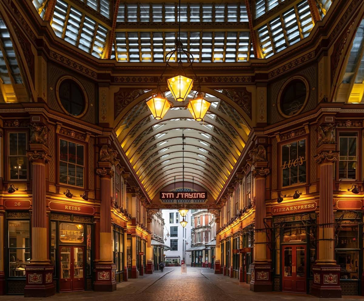 Vista em grande angular do interior ornamentado com teto de vidro do Leadenhall Market em Londres, com distintas colunas vitorianas, lanternas penduradas e arcos ricamente decorados.