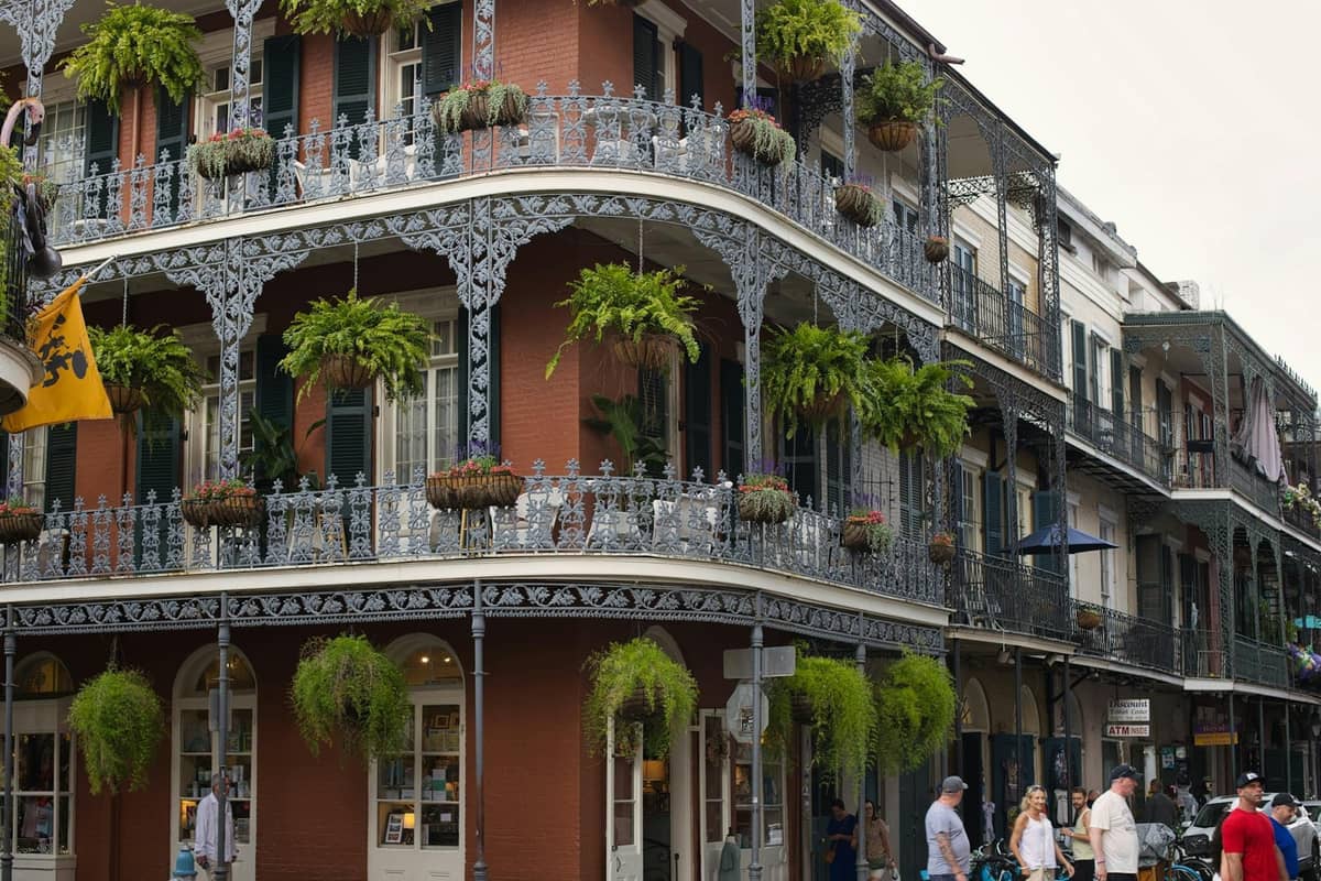 Ornate cast-iron balconies decorated with hanging plants on a classic brick building corner in the French Quarter, with pedestrians below.