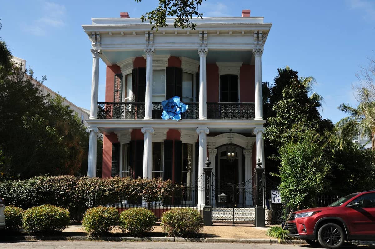 Two-story antebellum mansion with columns, wrought iron balcony, and lush greenery in front, capturing classic New Orleans architectural style.