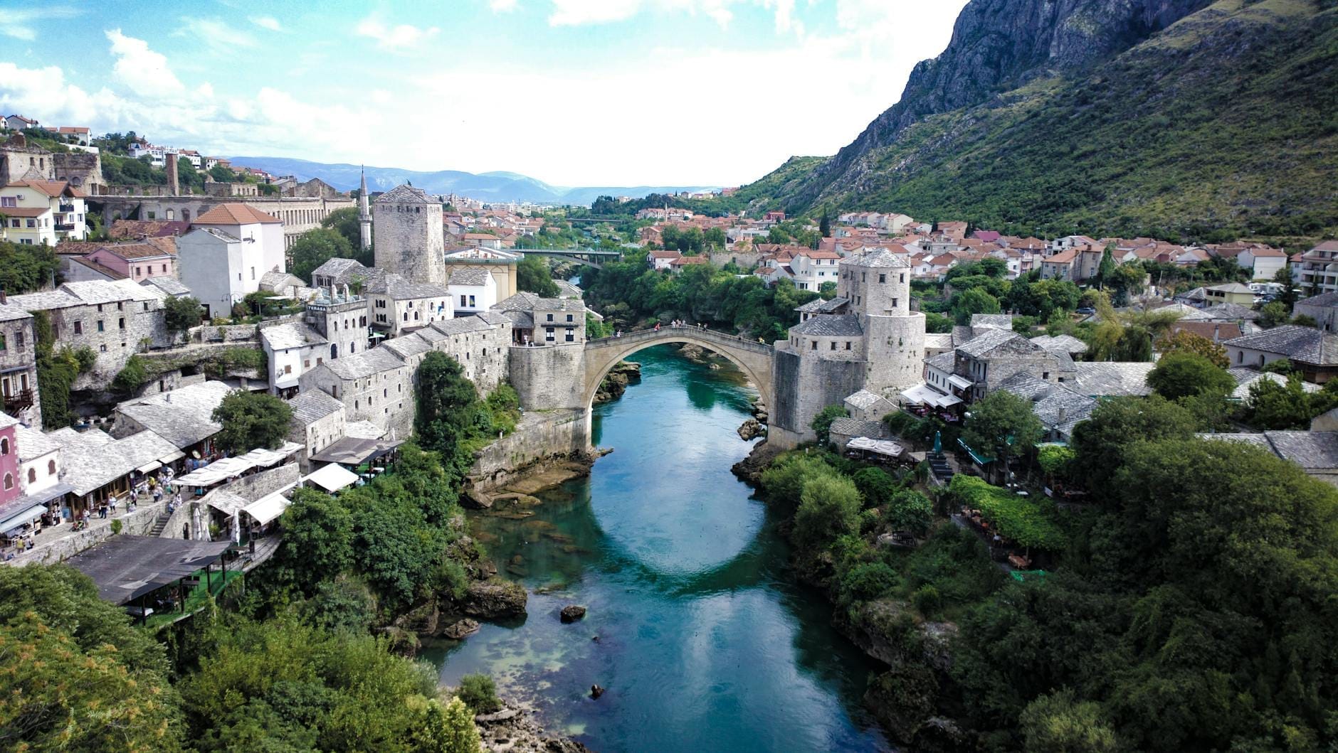 Vista aérea panorámica del Stari Most (Puente Viejo) sobre el esmeralda río Neretva en Mostar, rodeado de edificios históricos de piedra y montañas de intenso color verde.