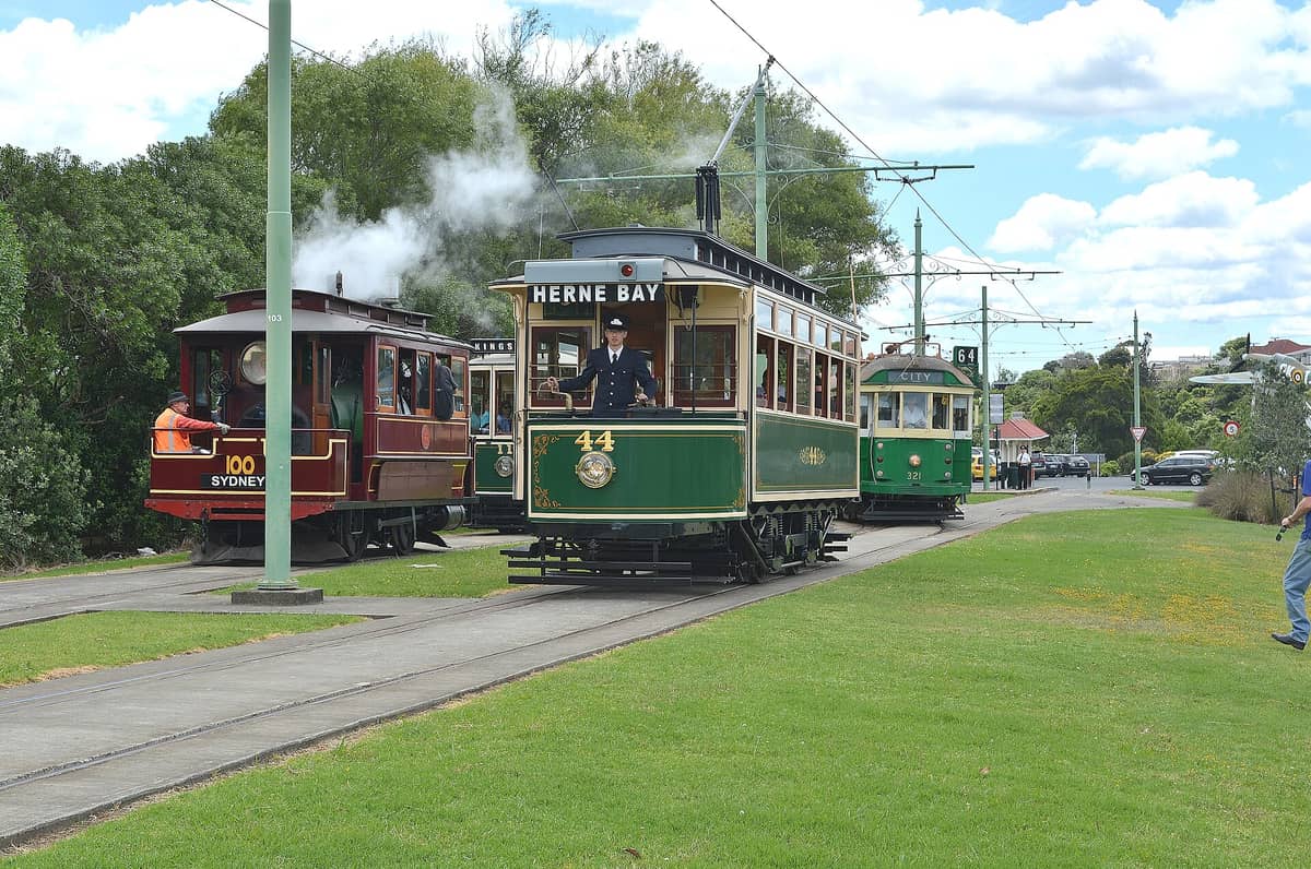 Drei historische Straßenbahnen, darunter eine grüne Herne-Bay-Tram, fahren auf Außengleisen im MOTAT, umgeben von Menschen und Grünflächen.