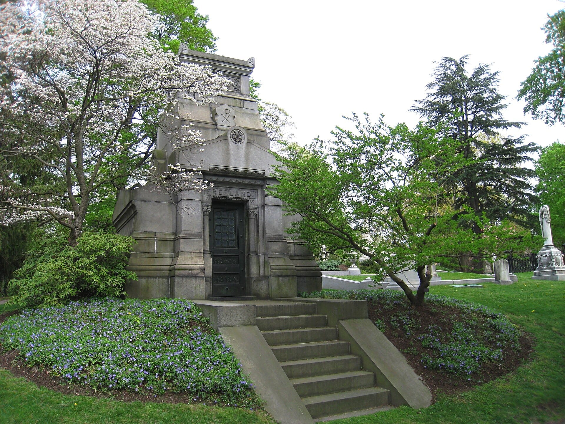 Mausoleo in pietra circondato da fiori primaverali, alberi rigogliosi e lapidi al Mount Auburn Cemetery in una giornata luminosa e serena.