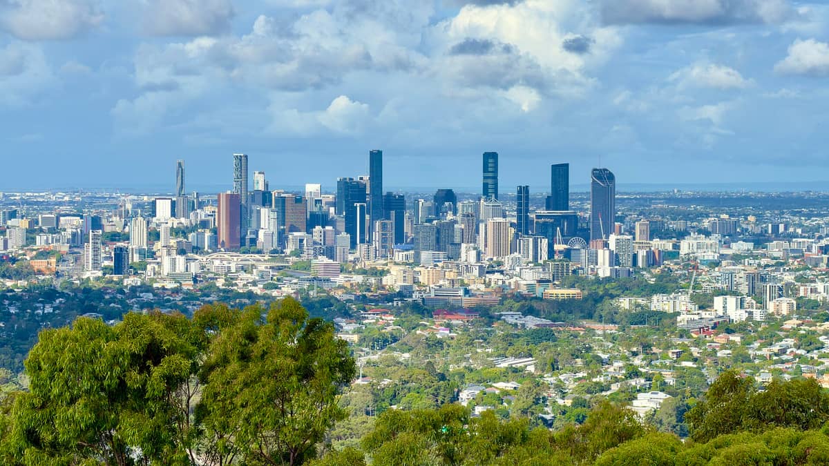 Panoramic view of Brisbane city skyline from Mount Coot-tha Lookout on a bright day, with tall buildings, suburban areas, and green trees in the foreground.