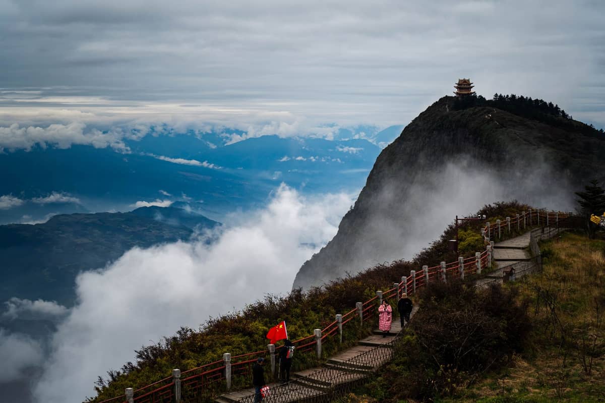 Dramatic summit view at Mount Emei with a temple pavilion on a high peak, clouds swirling below, and visitors hiking an ascending path.