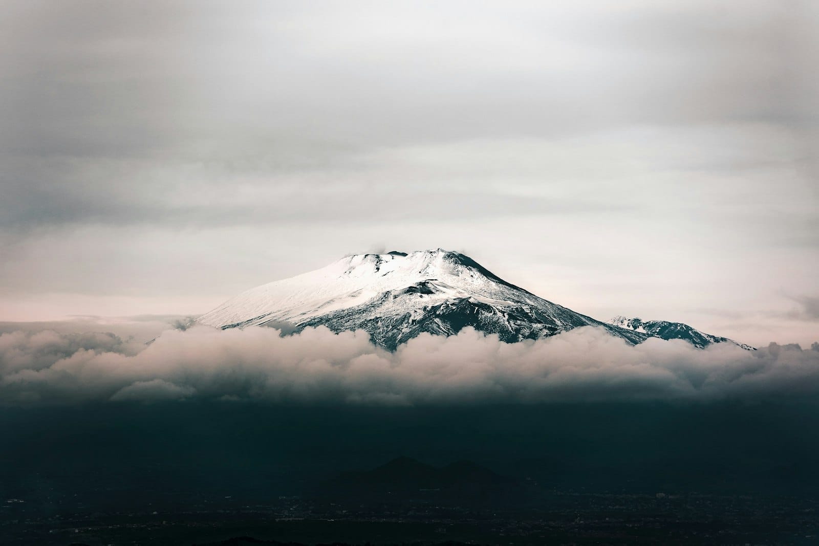 Der schneebedeckte Gipfel des Ätna erhebt sich über den Wolken, mit dramatischem Himmel und sichtbaren Vulkanhängen – ein markantes Wahrzeichen der sizilianischen Landschaft.