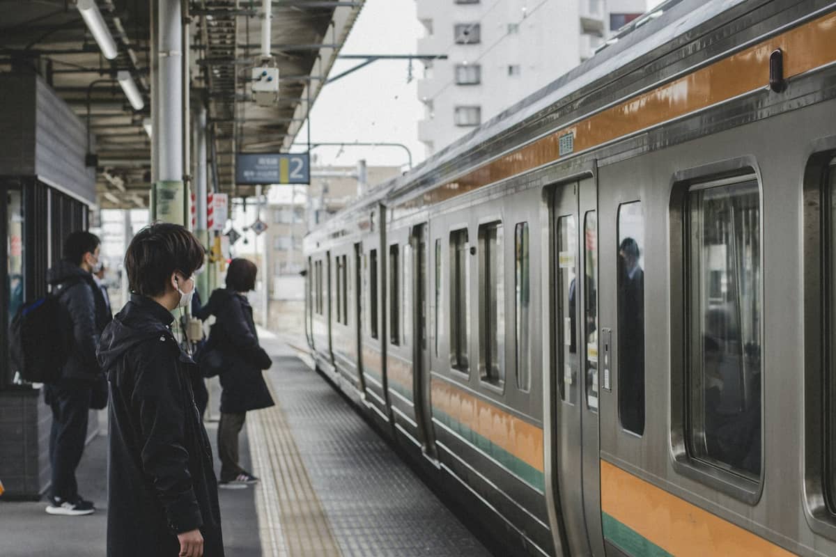 People waiting next to an orange-striped commuter train at a Japanese train platform with city buildings in the background.