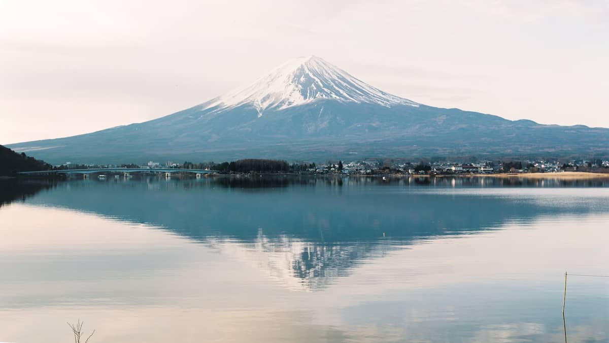 Snow-capped Mount Fuji reflected in the calm waters of Lake Kawaguchiko with a small lakeside town visible at the base.