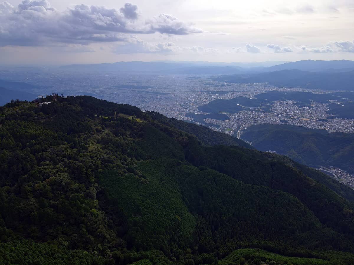 A sweeping aerial landscape of Mount Hiei with lush green forested ridges, temple structures near the summit, and the cityscape of Kyoto sprawling in the background under a cloudy sky.