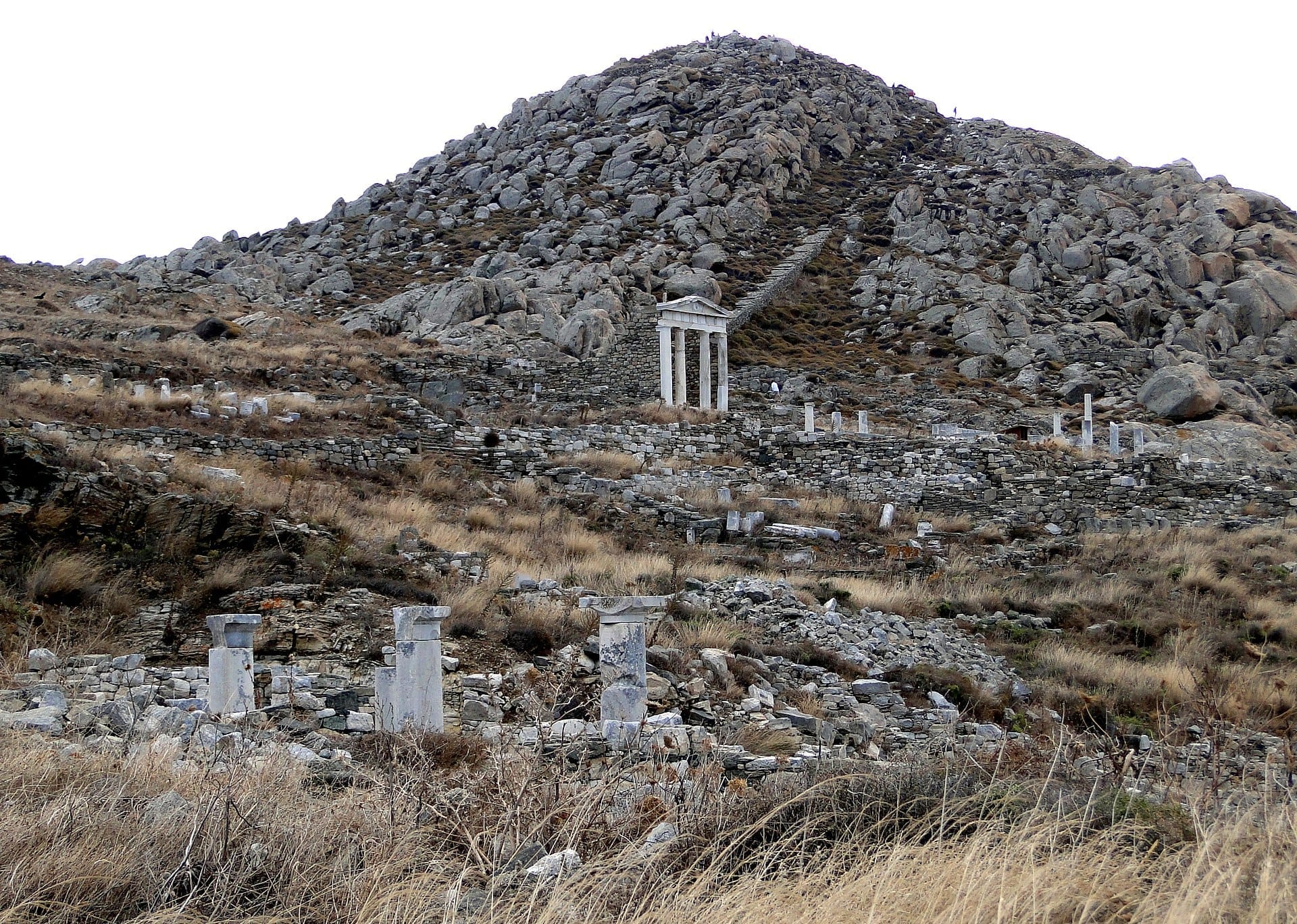 Laderas rocosas del Monte Kynthos en la isla de Delos con antiguas ruinas de piedra y un templo de estilo griego bajo un cielo nublado.