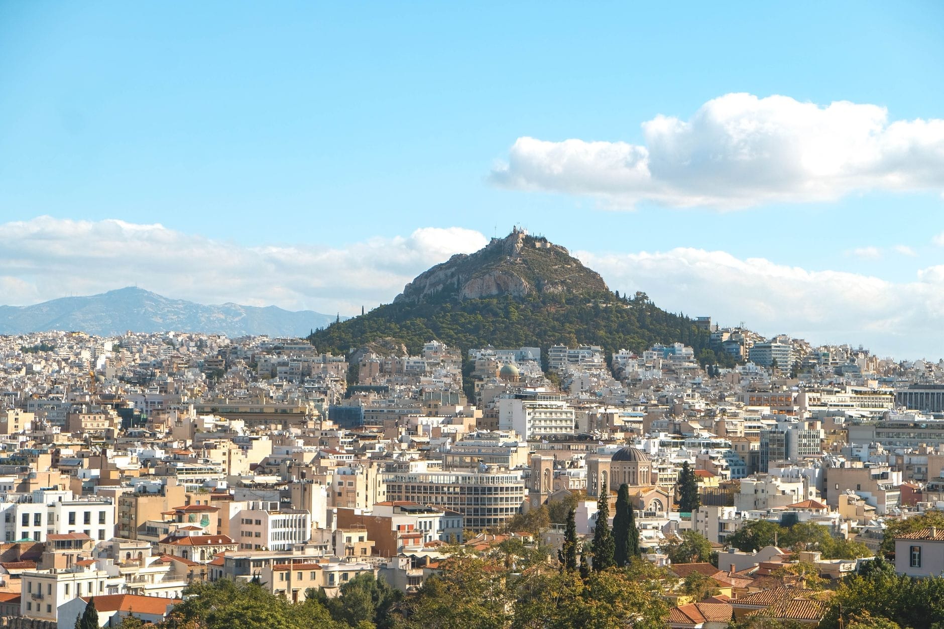 Il Monte Licabetto si erge sopra il panorama di Atene sotto un cielo azzurro brillante con nuvole leggere, offrendo una vista ampia e invitante per i viaggiatori.