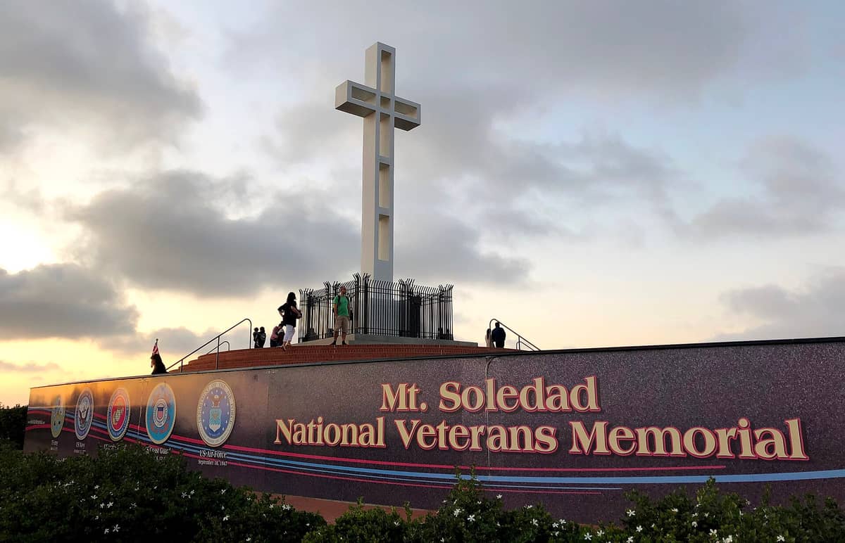 Das Mt. Soledad National Veterans Memorial bei Sonnenuntergang mit dem großen Betonkreuz auf dem Denkmal, dem offiziellen Schild und mehreren Besuchern.