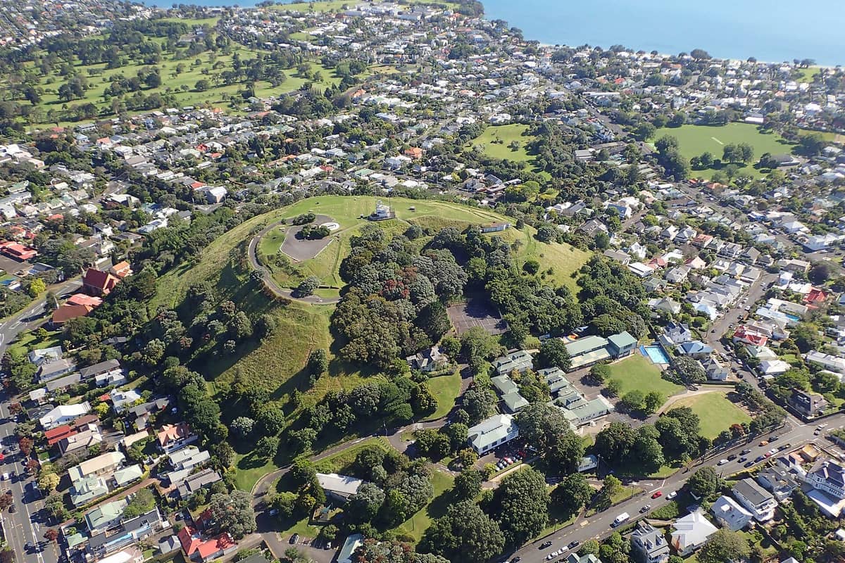 Luftaufnahme von Takarunga / Mount Victoria in Devonport mit grasbewachsenen Hängen, Gipfelstrukturen und umliegenden Wohngebieten bei klarem Tageslicht.