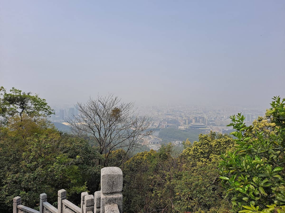View from Moxing Ridge summit showing lush trees in the foreground and Guangzhou city skyline partially obscured by haze.