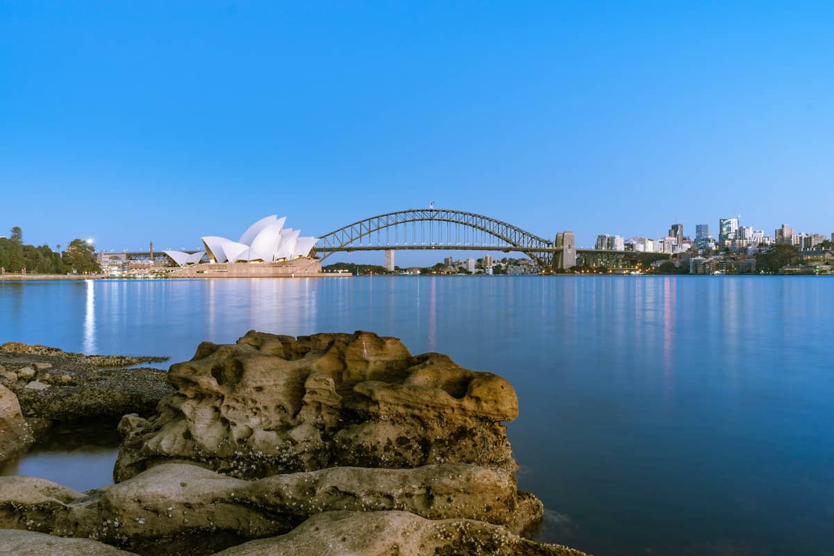 Vista panoramica dalla Sedia di Mrs Macquarie con rocce arenarie in primo piano e la Sydney Opera House e il Harbour Bridge perfettamente inquadrati oltre il porto al crepuscolo.