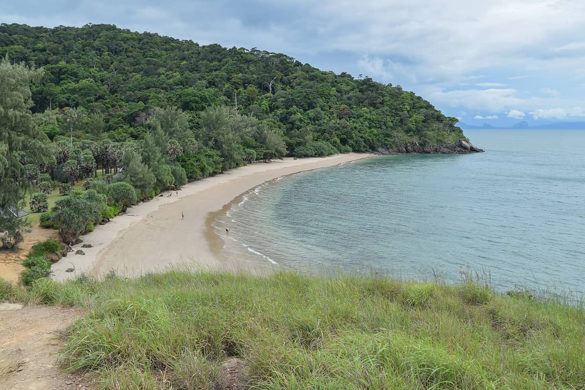 Ein geschwungener Sandstrand, umrahmt von dichtem grünem Wald im Mu Ko Lanta Nationalpark, aus der Vogelperspektive mit ruhigem Meer und wenigen Menschen am Strand.