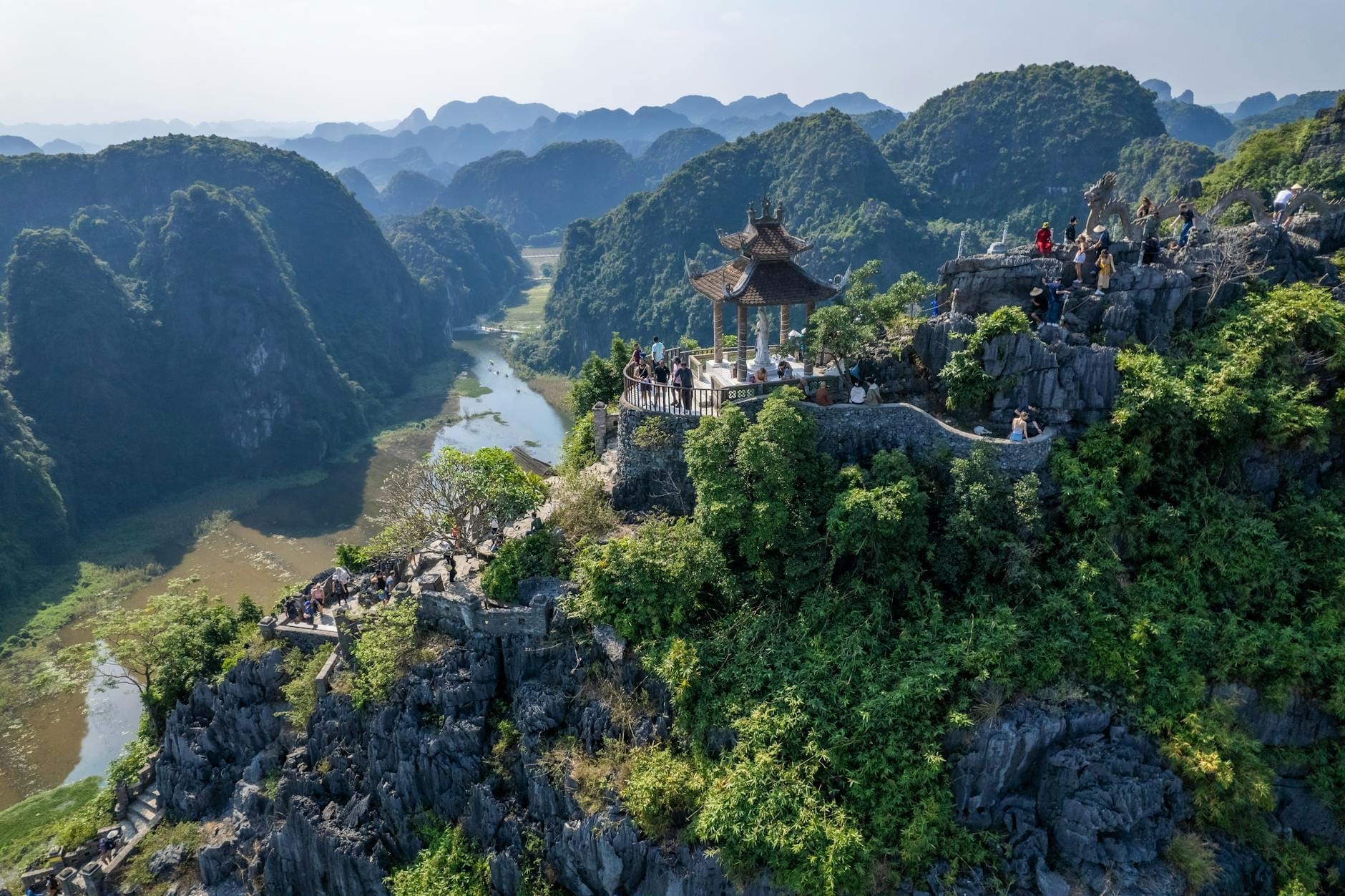 Aerial view of Hang Mua Peak with visitors at the summit pavilion, overlooking panoramic limestone mountains, winding river, and lush rice fields in Ninh Binh, Vietnam.
