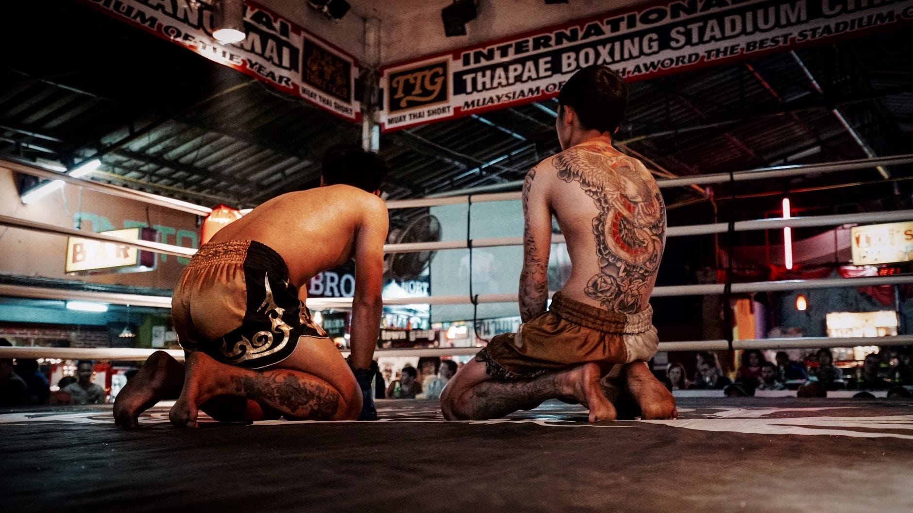 Dos luchadores de Muay Thai se arrodillan en un ring en el Thapae Boxing Stadium en Chiang Mai, rodeados por el público y la señalización del recinto.