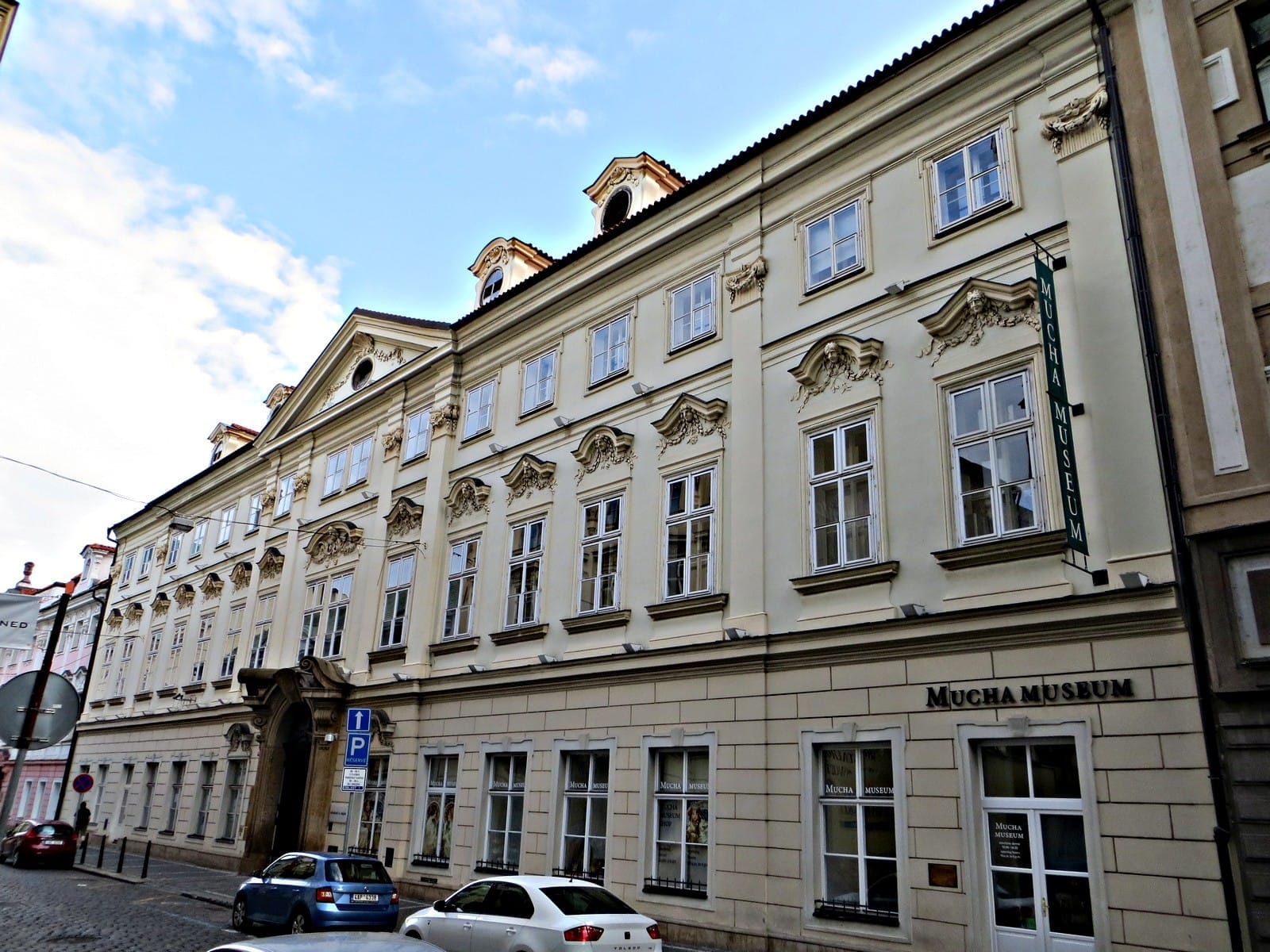 Außenansicht des Mucha Museums in Prag, untergebracht in einem prachtvollen cremefarbenen Palast aus dem 18. Jahrhundert mit verzierten Fenstern und klassischen architektonischen Details.