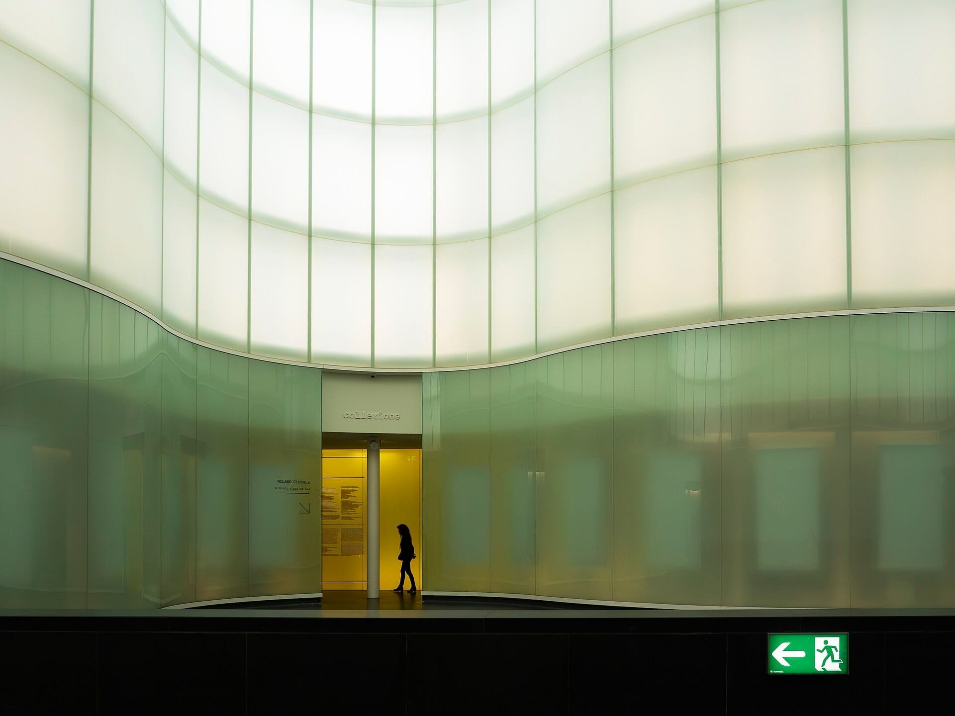 Vista interna del MUDEC – Museo delle Culture a Milano, con un visitatore solitario, morbide pareti in vetro curvate e una moderna illuminazione luminosa.