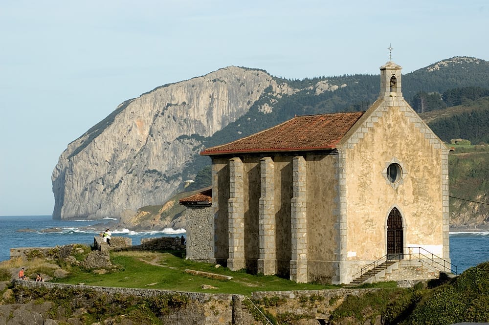 Steinkirche mit Blick auf das Meer in Mundaka, mit dramatischen Klippen und sanft geschwungenen grünen Hügeln im Hintergrund bei klarem Tageslicht.