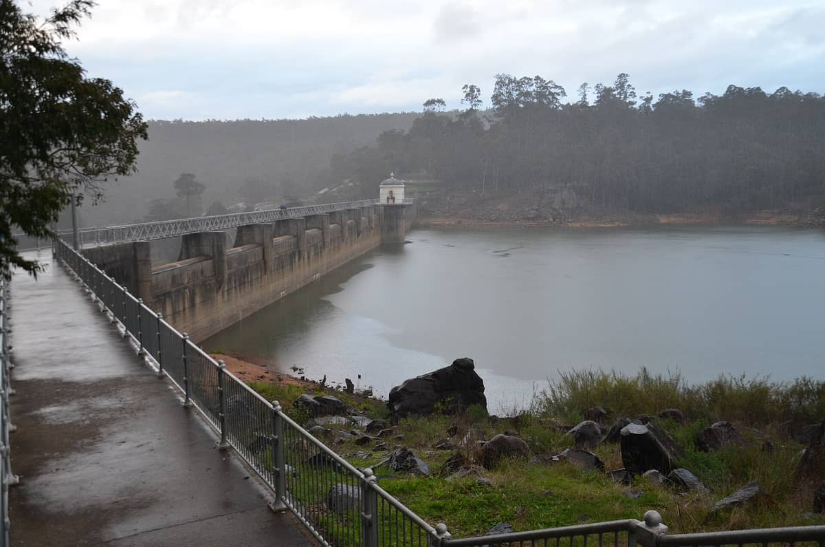Nasser Weg mit Metallgeländern führt zu Mundaring Weir, mit der Staumauer und bewaldeten Hügeln unter bedecktem Himmel.