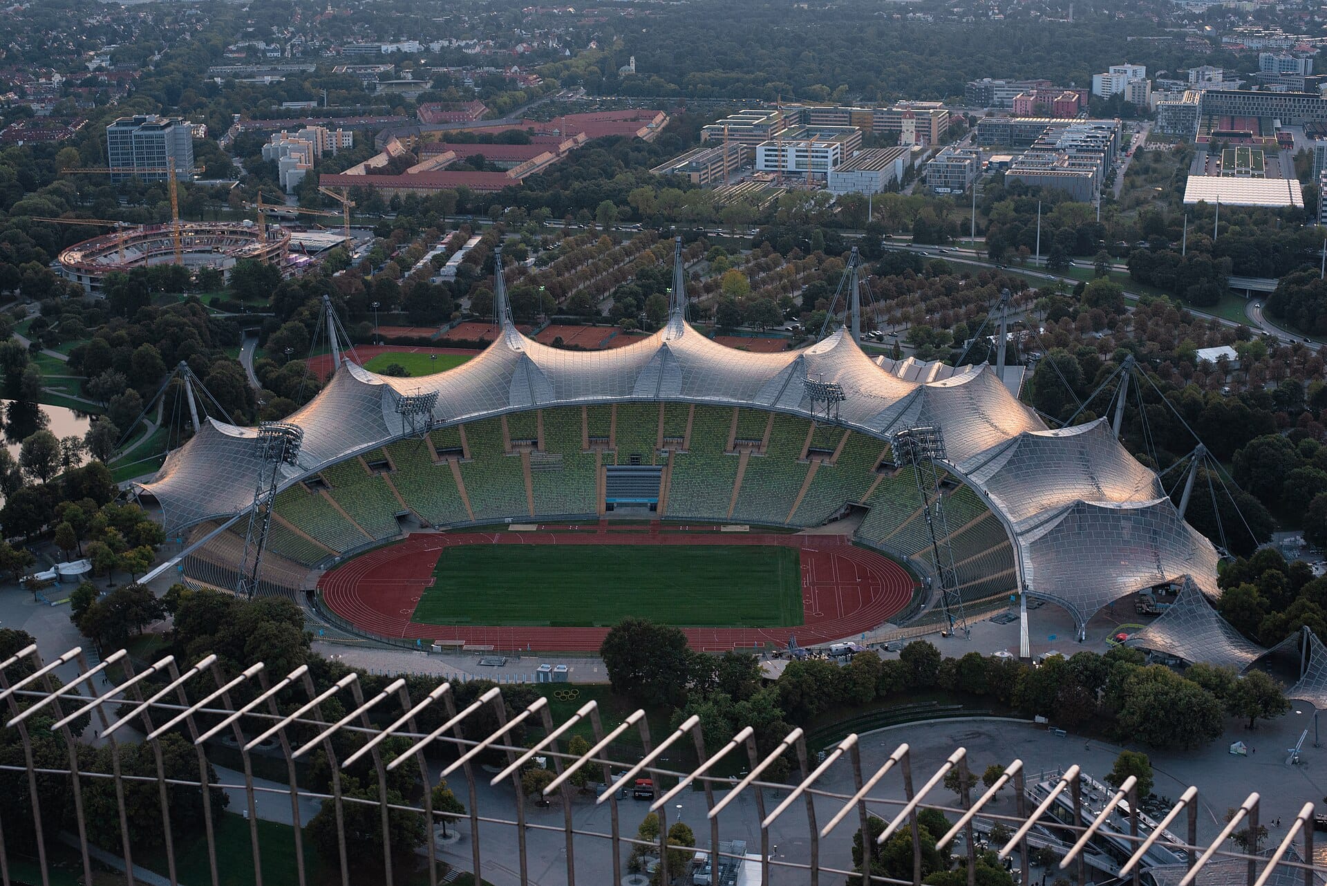 มุมมองทางอากาศของ Munich Olympiastadion ที่แสดงให้เห็นโครงสร้างหลังคาแบบ tensile โปร่งใสโค้งเว้าอันเป็นเอกลักษณ์ ล้อมรอบด้วย Olympic Park และทิวทัศน์เมือง
