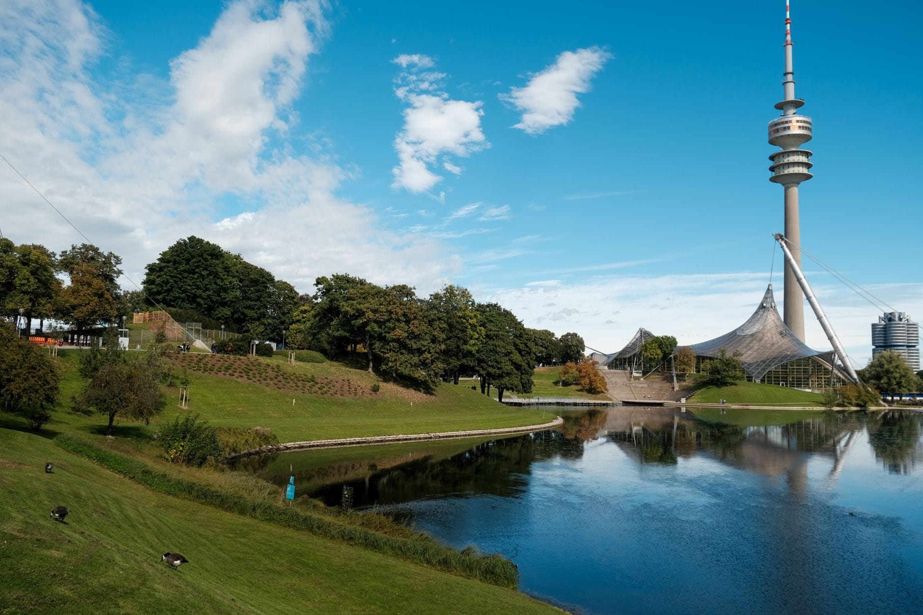 วิวของ Olympiapark ในมิวนิก พร้อม Olympic Tower อันโดดเด่น เนินเขาสีเขียวชอุ่ม และบึงน้ำสะท้อนแสงขนาดใหญ่ใต้ท้องฟ้าสีฟ้า