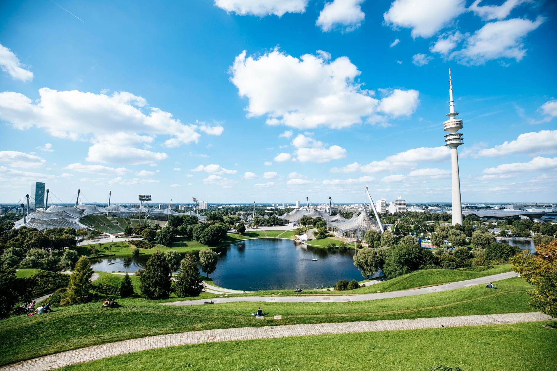 วิวกว้างของ Olympiapark München พร้อมทะเลสาบโอลิมปิก โครงสร้างหลังคาเต็นท์อันเป็นเอกลักษณ์ และ Olympic Tower ใต้ท้องฟ้าสีฟ้า