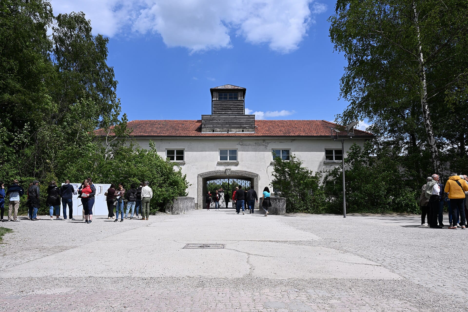 นักท่องเที่ยวรวมตัวกันอยู่หน้าประตูทางเข้าค่ายกักกัน Dachau ภายใต้ท้องฟ้าที่มีเมฆบางส่วน โดยมีต้นไม้และอาคารหลังคาสีแดงปรากฏให้เห็น